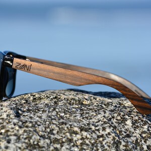 May include: Black sunglasses with wooden arms. The arms are dark brown wood with a natural grain pattern. The brand name "ZONI" is engraved on the right arm.