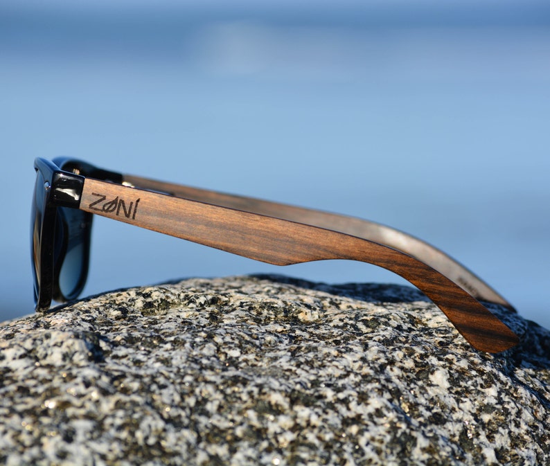 May include: Black sunglasses with wooden arms. The arms are dark brown wood with a natural grain pattern. The brand name "ZONI" is engraved on the right arm.