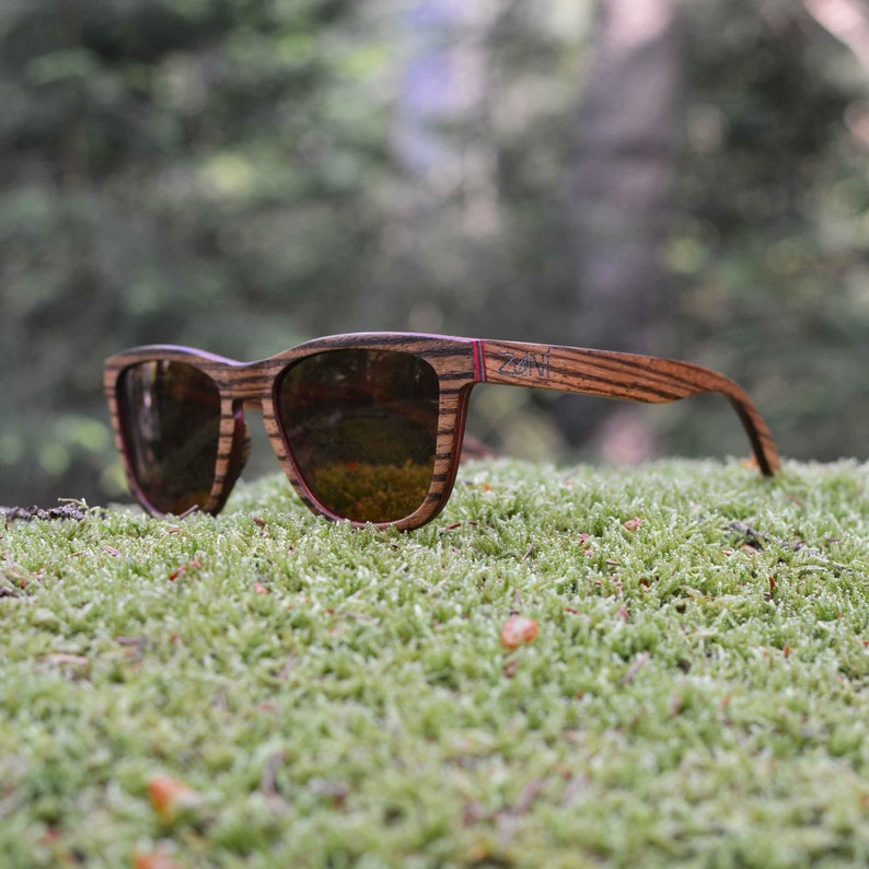 May include: Wooden sunglasses with brown and red frames and brown lenses. The sunglasses are resting on a bed of green moss.