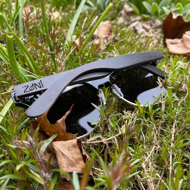 Puede incluir: Gafas de sol de madera negra con lentes negros y la palabra "ZONI" grabada en el lateral. Las gafas de sol est&aacute;n sobre un lecho de hierba verde.