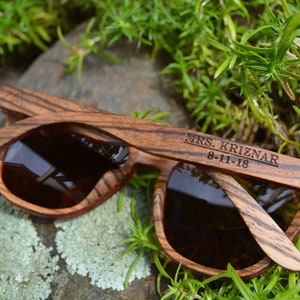 May include: Wooden sunglasses with brown lenses and a wood grain pattern. The sunglasses are engraved with the text "MRS. KRIZNAR 8-11-18".