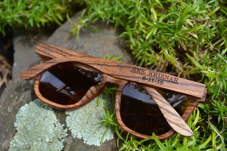May include: Wooden sunglasses with brown lenses and a wood grain pattern. The sunglasses are engraved with the text "MRS. KRIZNAR 8-11-18".