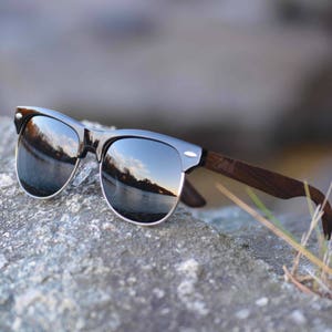 Puede incluir: Gafas de sol negras con lentes espejados y un marco de madera. Las gafas de sol est&aacute;n apoyadas en una roca gris.