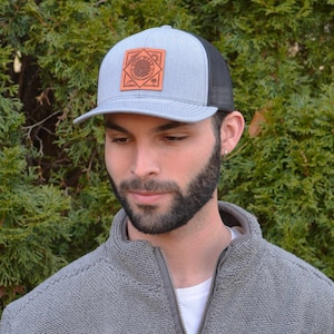 May include: A gray and black trucker hat with a brown leather patch featuring a sun, waves, and the text "Get Your Wave On".