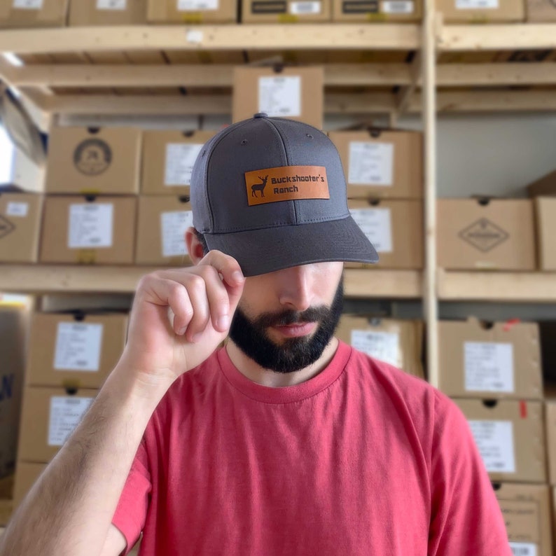 May include: A gray baseball cap with a brown leather patch that says "Buckshooter's Ranch" and a silhouette of an animal. The person wearing the cap is wearing a red t-shirt.