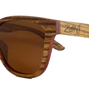 May include: Brown wood sunglasses with brown lenses and a red stripe on the side. The sunglasses have the word "ZONI" engraved on the side.