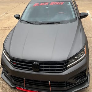 May include: A gray Volkswagen Passat with a black grille and a red "BLACK ROUX" windshield banner. The car has a black front splitter with a red lip.