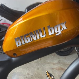 May include: A close-up of a gold-colored electric bicycle with a black frame and a black seat. The bicycle has a black pedal and a white sticker that says "BIGNIU bgX".