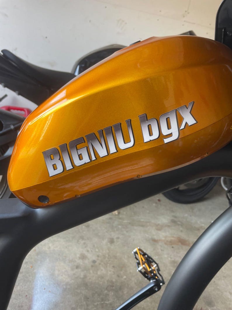 May include: A close-up of a gold-colored electric bicycle with a black frame and a black seat. The bicycle has a black pedal and a white sticker that says "BIGNIU bgX".