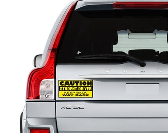 Könnte beinhalten: Ein gelbes und schwarzes Warnschild auf der Rückseite eines silbernen Autos mit der Aufschrift "CAUTION STUDENT DRIVER KEEP BACK WAY BACK".