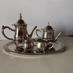 May include: A silver-plated tea service set consisting of a large teapot, a smaller teapot, a lidded sugar bowl, and a creamer, all arranged on an oval serving tray. Each piece features ornate, curved handles and decorative feet with polished surfaces.