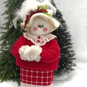 May include: A white snowman wearing a red and white plaid skirt, a red sweater, a white crocheted collar, and a white hat with a white feather trim. The snowman has rosy cheeks and black eyes.