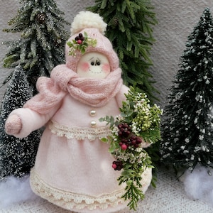 May include: A pink snowman doll wearing a pink knitted hat with a white pom pom, a pink scarf, and a pink dress with white lace trim. The snowman is holding a bouquet of green and white flowers with red berries.