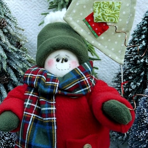 May include: A snowman doll wearing a red coat, green hat, and a plaid scarf. The snowman is holding a green and white kite with red and white polka dots.