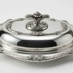 May include: A polished silver-colored oval serving dish with a matching lid. The lid features a central decorative handle and an embossed floral border. The base tray has a wide, scalloped rim with intricate scrollwork and floral detailing.