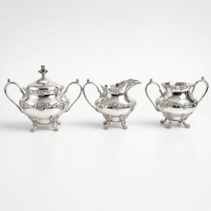 May include: A three-piece silver tea service set consisting of a lidded sugar bowl, a creamer, and a waste bowl. Each piece features ornate floral embossed detailing, curved handles, and stands on four small, decorative feet with a polished finish.