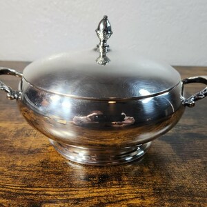 以下が含まれることがあります： 磨き上げられた銀色の金属製サービングボウル。蓋と2つの装飾的なサイドハンドルが付いています。蓋の中央には小さな装飾的なつまみが付いています。ボウルは滑らかで反射する表面を持ち、円形の台座の上に置かれています。