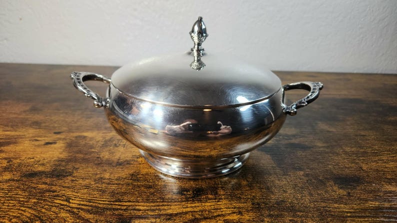以下が含まれることがあります： 磨き上げられた銀色の金属製サービングボウル。蓋と2つの装飾的なサイドハンドルが付いています。蓋の中央には小さな装飾的なつまみが付いています。ボウルは滑らかで反射する表面を持ち、円形の台座の上に置かれています。