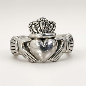 May include: A silver Claddagh ring featuring a central heart shape held by two hands, topped with a detailed crown. The metal has a polished finish with darkened recessed areas to highlight the intricate textures of the crown and cuffs.