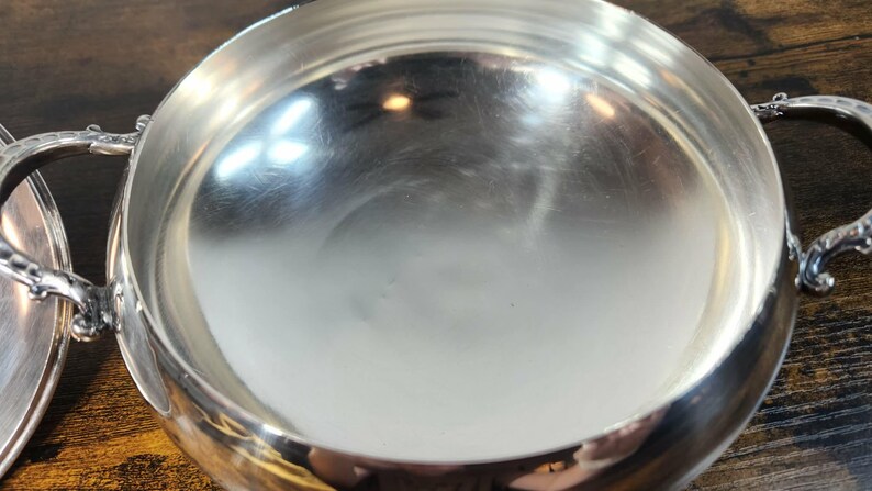 以下が含まれることがあります： 光沢のある銀色の金属製サービングボウル。内側は滑らかで反射する表面になっており、両サイドには装飾が施された曲線的なハンドルが付いています。ボウルは幅の広い円形の縁と深い容器で構成され、全体に金属的な仕上げが施されています。