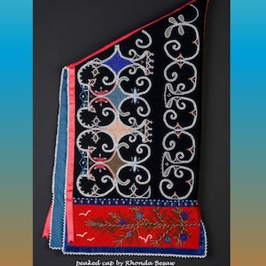 May include: A black peaked cap with intricate beadwork. The cap features white, blue, and brown beads in a repeating pattern. A red band with a detailed design and the text "peaked cap by Rhonda Besaw" is at the bottom. The year 2022 is also visible.