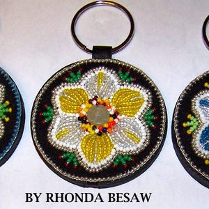 Pode incluir: Três chaveiros redondos com desenhos florais de contas. Cada chaveiro apresenta um verso de couro preto, um chaveiro prateado e um desenho floral central em vermelho, amarelo e azul. O texto "BY RHONDA BESAW" está na parte inferior.