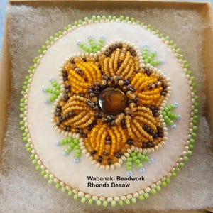 May include: A white felt circle with a beaded flower design in shades of brown, yellow, and green. The flower has a brown center with a clear, iridescent stone. The text "Wabanaki Beadwork Rhonda Besaw" is printed below the flower.