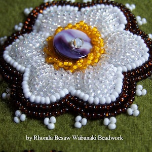 May include: Close-up of a beaded flower with a purple and white shell button in the center. The flower is surrounded by brown beads and white beads. The background is green felt. "by Rhonda Besaw Wabanaki Beadwork" is written below the flower.