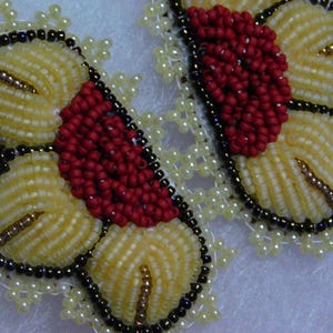 May include: Close-up of two beaded butterfly earrings. The earrings feature a combination of yellow, red, and black beads. The wings are yellow with red centers, outlined in black beads, and accented with gold-colored beads. The earrings are surrounded by small, white beads.