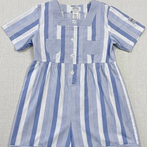 May include: A blue and white striped romper with short sleeves and a square neckline. The romper has two small pockets and buttons down the front. The fabric appears to be cotton. The garment is designed for children.