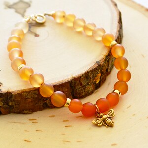 May include: A honey-colored and orange gemstone bracelet with a gold bee charm. The bracelet is made of round beads and has a gold clasp.