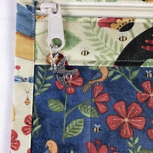 Puede incluir: Primer plano de un art&iacute;culo de tela con cremallera y un charm de p&aacute;jaro. La tela presenta un estampado floral y de p&aacute;jaros en colores azul, rojo y crema. La cremallera es blanca y el charm es plateado.