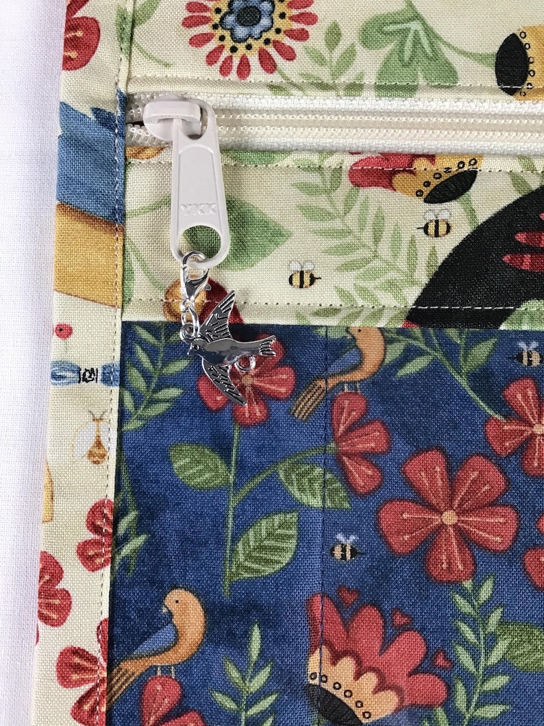 Puede incluir: Primer plano de un art&iacute;culo de tela con cremallera y un charm de p&aacute;jaro. La tela presenta un estampado floral y de p&aacute;jaros en colores azul, rojo y crema. La cremallera es blanca y el charm es plateado.