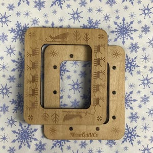 May include: Two wooden embroidery hoops with decorative laser-cut designs. The hoops are light brown and feature a rectangular shape with rounded corners. The text "MesaQuillCo" is visible on the bottom hoop. The background is a white fabric with blue snowflake patterns.