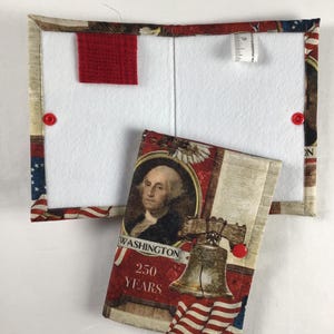 May include: A patriotic fabric notebook cover and matching small notebook. The cover features a red, white, and blue American flag design with a portrait of George Washington and the text "250 YEARS". The cover has a red felt pocket and red button closures.