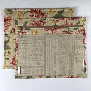 May include: Two beige quilted project bags with a floral pattern in red, yellow, and green. One bag is open, revealing a beige fabric panel with cross-stitch alphabet and number charts. The bags have a zipper closure.