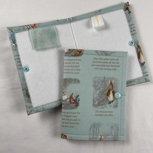 May include: A light blue fabric book cover and matching bookmark with illustrations of rabbits and text from a children's story. The cover has a button closure and a small felt square. The bookmark has a small loop.