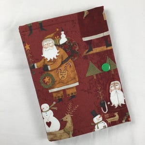 May include: A rectangular fabric cover featuring a dark red background with a repeating pattern of Santa Claus figures, snowmen, reindeer, and evergreen trees. A small green plastic snap button secures the front flap of the cover.
