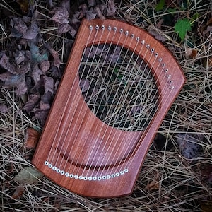 May include: A wooden lyre, a stringed musical instrument, with a curved design and numerous strings. The lyre is crafted from reddish-brown wood and features tuning pegs. It rests on a bed of dried leaves and grass, creating a natural setting.