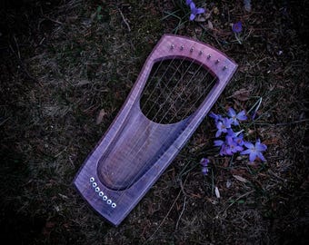 May include: A purple and brown wooden lyre with strings, lying on a bed of green grass and purple flowers.