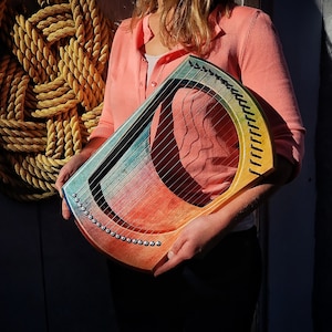 Puede incluir: Un laúd de madera de color arcoíris con cuerdas plateadas. El laúd lo sostiene una persona que lleva una camisa rosa.