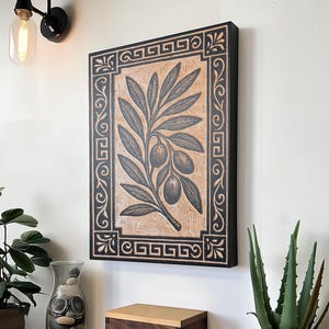 Peut inclure: Tableau encadré représentant une branche d'olivier avec un cadre noir et un fond beige. La branche d'olivier est illustrée en gris foncé. L'œuvre est exposée au-dessus d'une boîte en bois et de plantes en pot.
