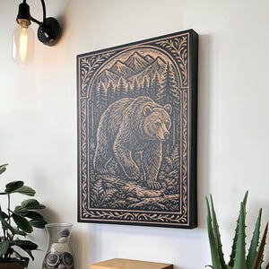May include: A framed woodcut art piece featuring a detailed bear illustration in a forest setting. The artwork is in a black frame and is mounted on a white wall. The bear is brown and the forest is detailed with trees and mountains.