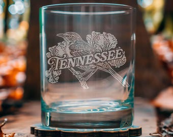 Peut inclure: Un verre transparent avec un motif gravé représentant un oiseau perché sur une branche, avec le mot "Tennessee" en dessous. Le verre est posé sur un dessous de verre en bois, avec un arrière-plan flou de forêt.