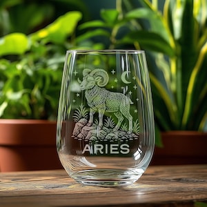 May include: A clear stemless wine glass featuring an etched design of a ram standing on a rocky outcrop, surrounded by small stars and a crescent moon. The word ARIES is etched in capital letters below the ram illustration on the glass surface.