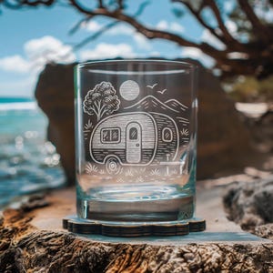 Puede incluir: Un vaso transparente con un diseño grabado de una caravana vintage, un árbol, el sol, montañas y pájaros. El vaso está sobre un posavasos de madera, con un fondo borroso del océano y el cielo.