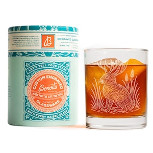 May include: A custom engraved glass with a rabbit design, next to a matching teal and orange gift box. The glass is filled with a brown liquid and an ice cube. The box reads "Let's tell your story" and "Custom Engraved Glassware".