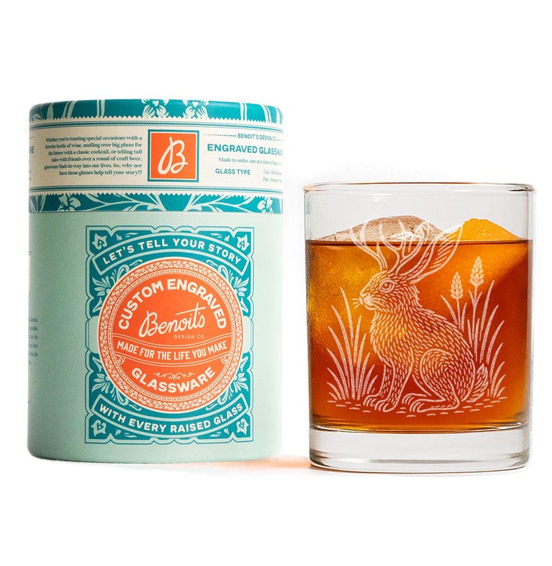 May include: A custom engraved glass with a rabbit design, next to a matching teal and orange gift box. The glass is filled with a brown liquid and an ice cube. The box reads "Let's tell your story" and "Custom Engraved Glassware".