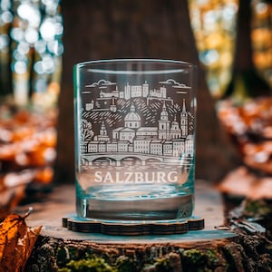 May include: A clear glass tumbler with a detailed white etched design of the Salzburg skyline, including buildings, a bridge, and the word "SALZBURG." The glass sits on a wooden coaster, with a blurred natural background.