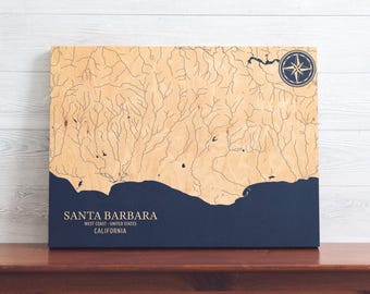 May include: A wooden map of Santa Barbara, California, with blue rivers and a blue ocean. The map is on a wooden background and has a compass rose in the top right corner. The text "SANTA BARBARA" is printed below the map, along with "WEST COAST - UNITED STATES" and "CALIFORNIA".